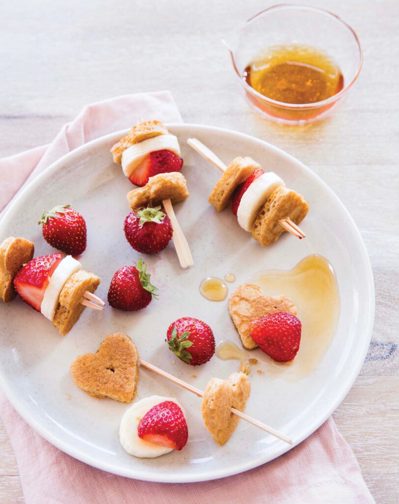 mini skewers with pancake, banana and strawberry on a white plate with syrup in a glass container on the side 