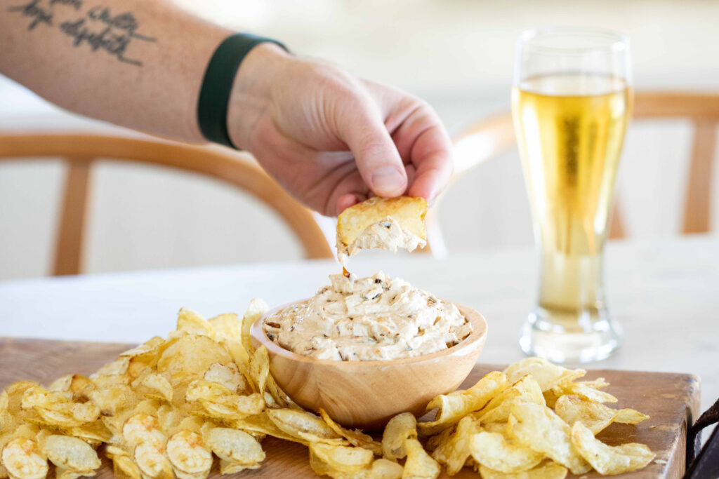 A male hand with a tattoos on the forearm dips a potato chip into the dairy-free creamy onion dip