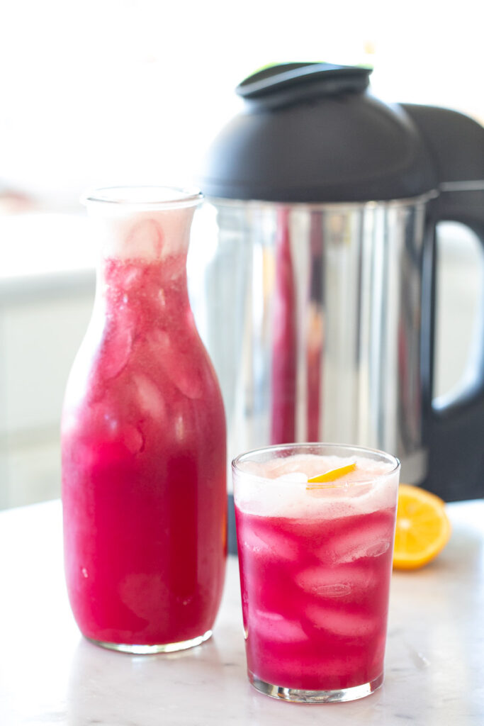 A pitcher and glass of Wild Blueberry Lavender Lemonade sits on a counter top with the Almond Cow in the background