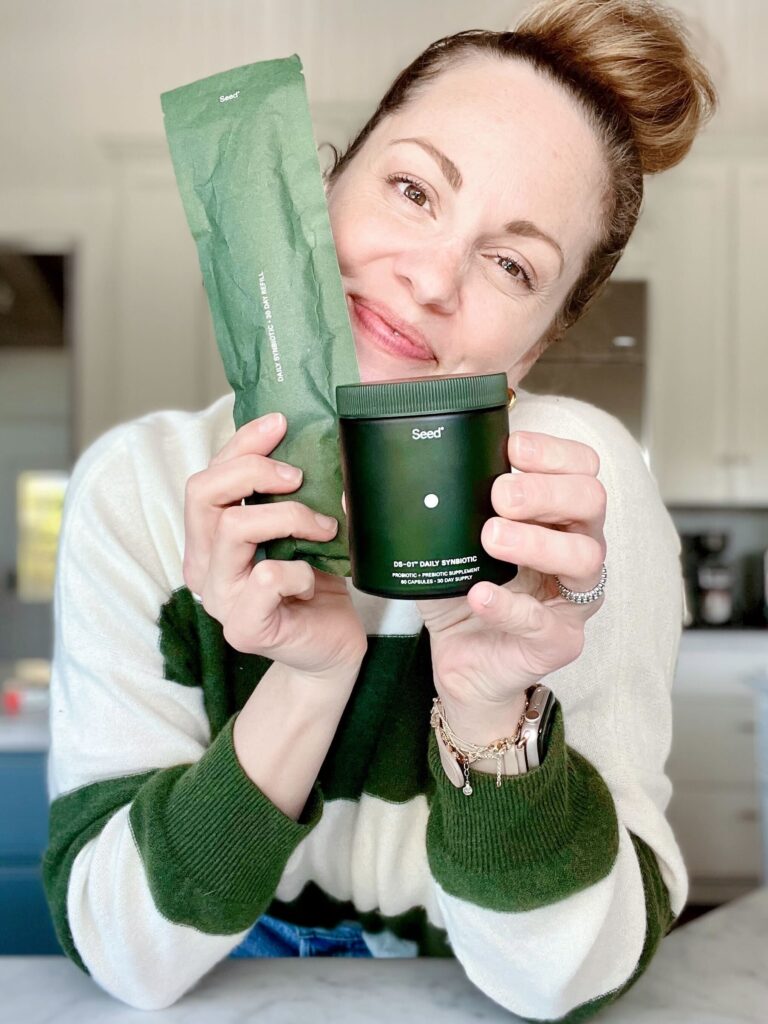 woman wearing a green and white striped sweater poses smiling at the camera while holding Daily Synbiotic