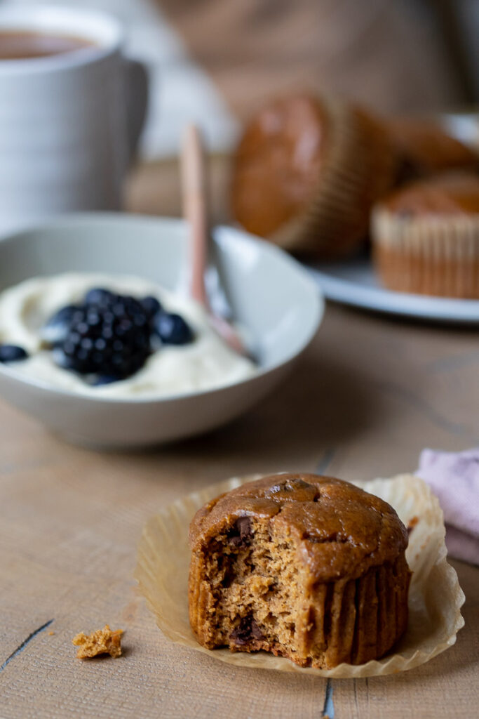 Gluten-Free Pumpkin Muffins on a wooden table, one is bitten and the rest are on a platter in the background next to a bowl of yogurt and fresh berries