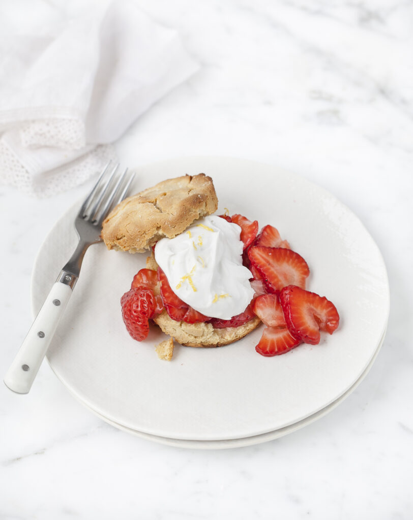 Gluten-Free Strawberry Shortcake on a white plate sitting on a white counter. A napkin lays next to the plate and a silver fork with white handle rests on the plate.