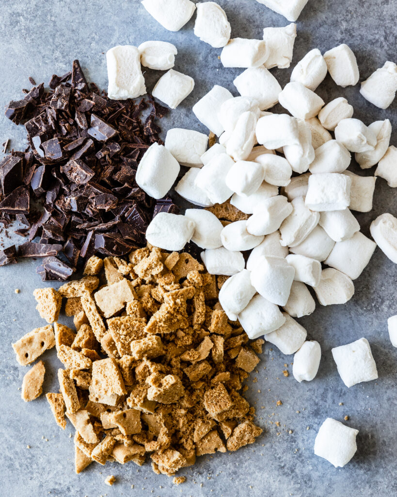 chocolate chunks, broken up pieces of graham crackers, and pieces of marshmallow lay together on a gray counter