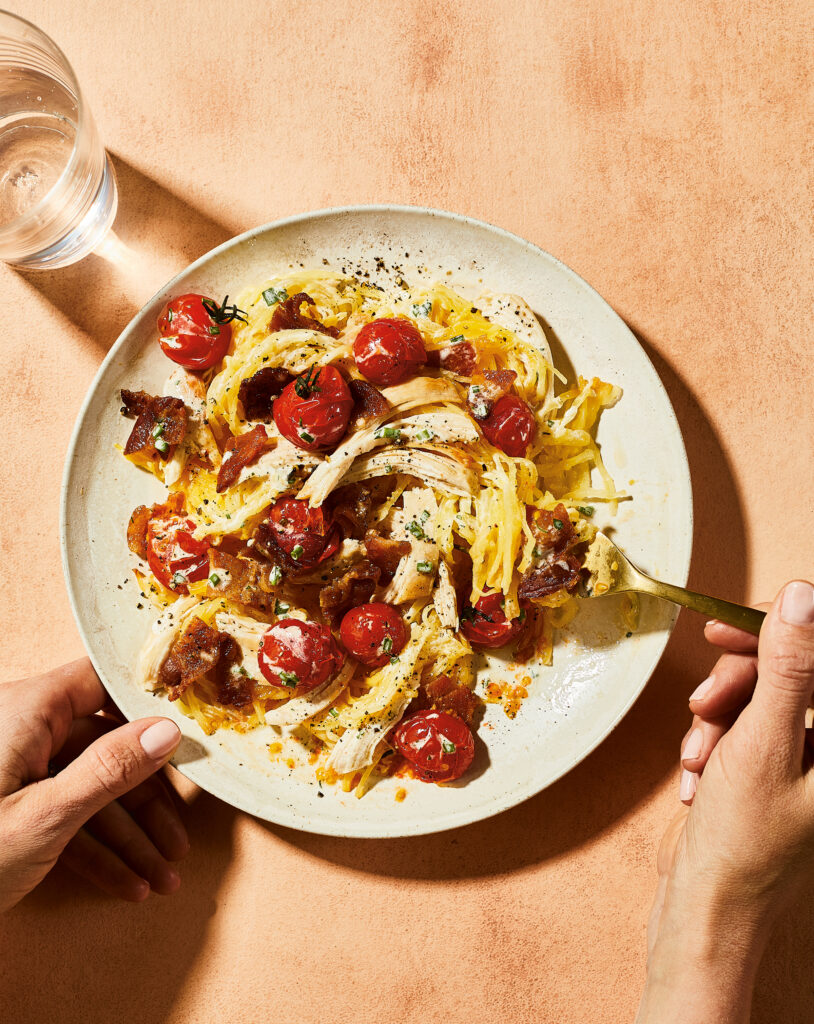 Chicken Bacon & Ranch Squash Noodles on a plate with hands pictured holding it and utilizing a fork. A glass of water sits nearby. 