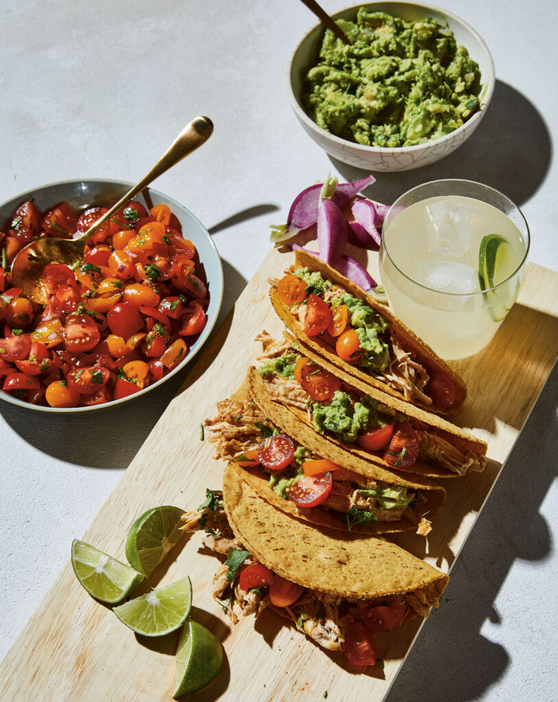 Paleo Salsa Chicken Tacos displayed on a wooden cutting board - a margarita, a bowl of guacamole and a bowl of chopped tomatoes are displayed with the tacos