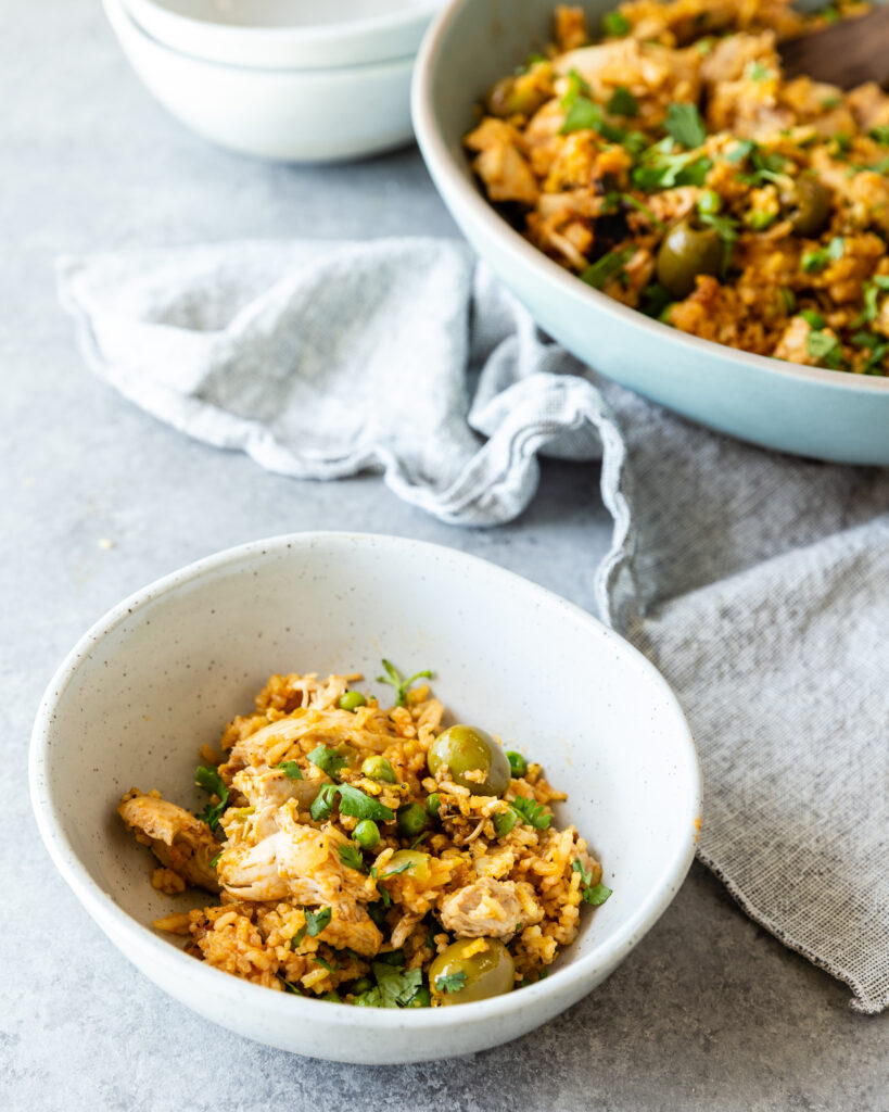 Instant Pot Arroz con Pollo served in a white bowl that is next to a kitchen towel. Instant Pot Arroz con Pollo is in a larger serving platter in the background. 