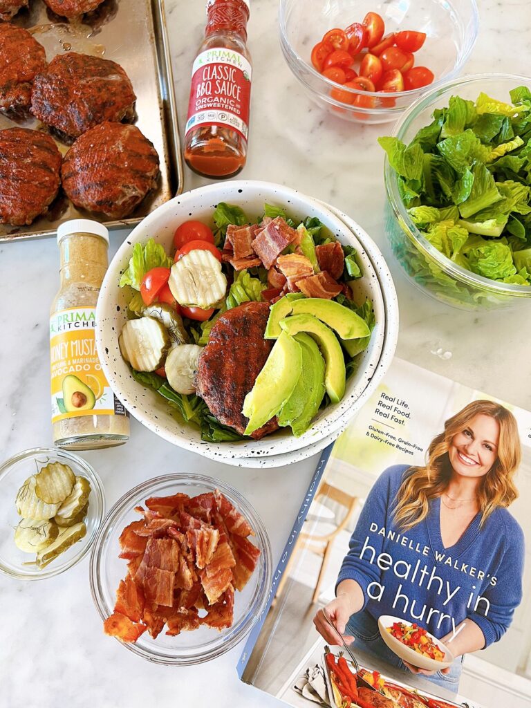 Paleo BBQ Bacon Burger Bowls on counter with the required ingredients surrounding the bowl with a copy of Healthy In a Hurry and Primal Kitchen sauces displayed