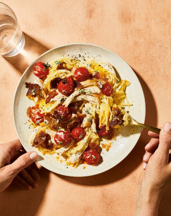 A medley of Chicken Bacon Ranch Squash noodles are pictured on a white platter with one hand holding the plate and the other holding a fork. A glass of water rests on the table to the left of the plate. 