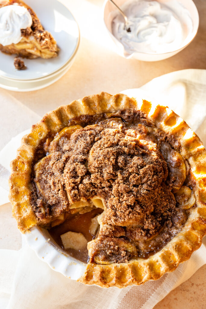 Gluten-Free Dutch Apple Pie with a golden crust, with one slice on a small plate and a bowl of coconut whipped cream next to it 