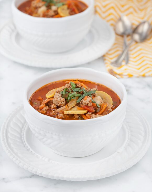 Paleo Minestrone is served in a white bowl atop of a white platter. This rests on a marble counter and in the background you can see another bowl of minestrone and 2 silver spoons on a white and yellow chevron napkin