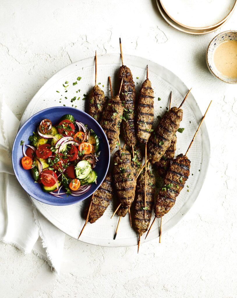 Kebabs resting on a white plate with a tomato medley in a small blue bowl next to it 