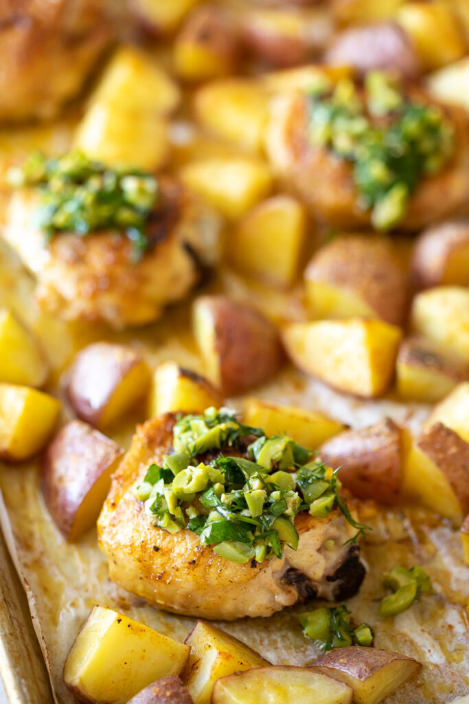 Close up image of roasted chicken thighs and potatoes on a sheet pan with lemon olive salsa on top.