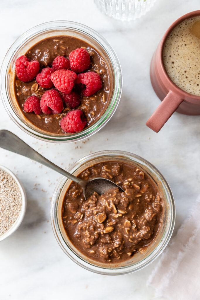 image from above of chocolate protein overnight oats in a glass bowl with a spoon next to a bowl of raspberries