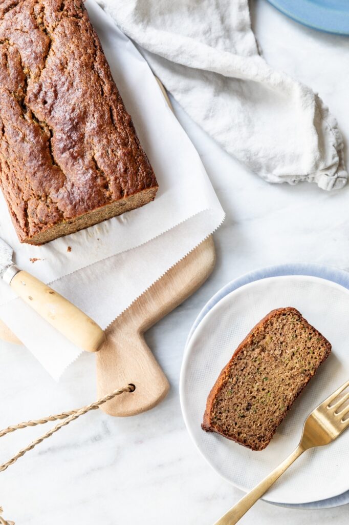 Above shot of baked almond flour zucchini bread on a wooden cutting board and a slice of bread on a white plate with a golden fork